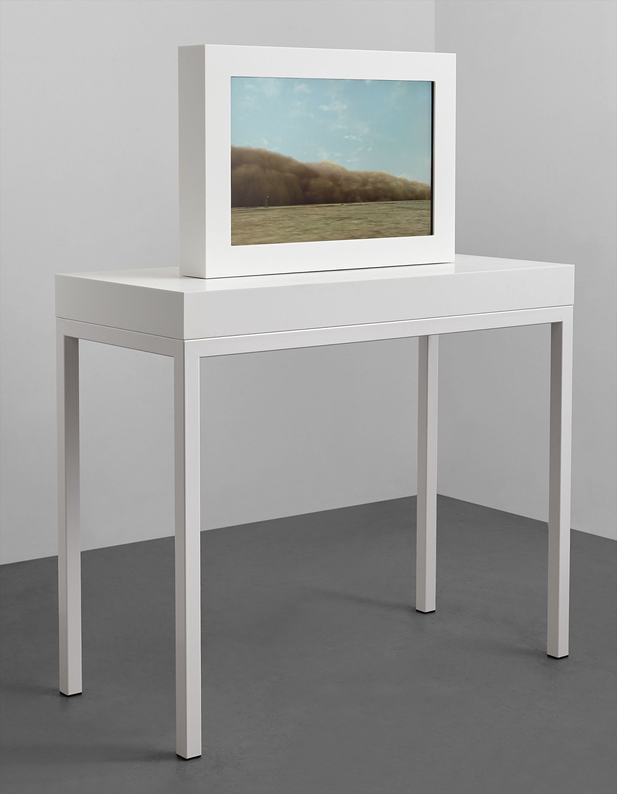 A white framed screen on a white table showing a brown dust cloud rolling over a grassy plain
