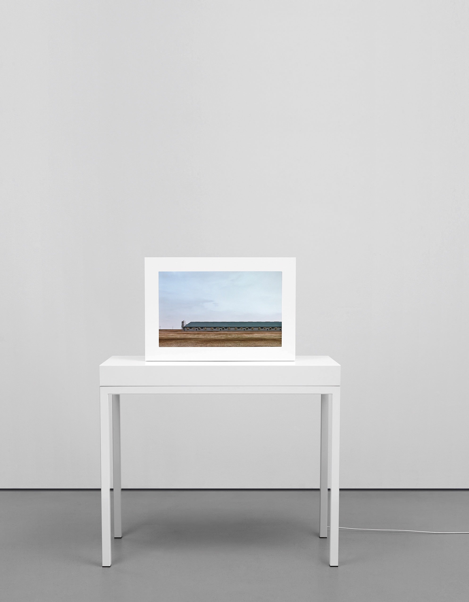 A white framed screen on a white table showing a long grey farm shed on flat brown land